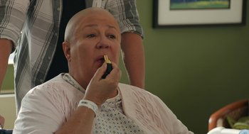 Movie still from “The Hollars” (2016), directed by John Krasinski – An older bald woman eating a banana while sitting in a chair; Close Up shot, Over the shoulder angle