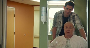 Movie still from “The Hollars” (2016), directed by John Krasinski – A man standing next to an older woman in a hospital room; Medium shot, Over the shoulder angle