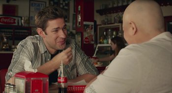 Movie still from “The Hollars” (2016), directed by John Krasinski – A man sitting at a table with a bottle of soda; Medium shot, Over the shoulder angle