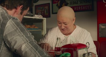 Movie still from “The Hollars” (2016), directed by John Krasinski – A bald woman sitting at a table with a bottle of soda; Close Up shot, High angle