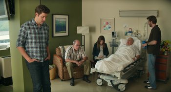 Movie still from “The Hollars” (2016), directed by John Krasinski – A man sitting in a chair in front of an older man in a hospital bed; Wide shot, Over the shoulder angle