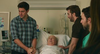 Movie still from “The Hollars” (2016), directed by John Krasinski – Three men standing next to an older man in a hospital bed; Medium shot, Over the shoulder angle