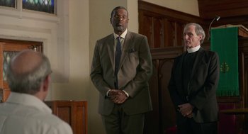 Movie still from “The Hollars” (2016), directed by John Krasinski – A man in a suit standing in front of two other men; Medium shot, Over the shoulder angle