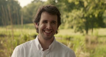 Movie still from “The Hollars” (2016), directed by John Krasinski – A man with long hair and a white shirt smiling for the camera; Close Up shot, Over the shoulder angle