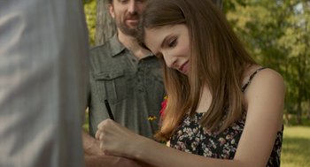 Movie still from “The Hollars” (2016), directed by John Krasinski – A woman is writing on a piece of paper while standing next to a man; Medium shot, Over the shoulder angle