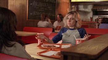 Movie still from “The House of the Devil” (2009), directed by Ti West – A woman sitting at a table with a plate of pizza; Medium shot, Over the shoulder angle