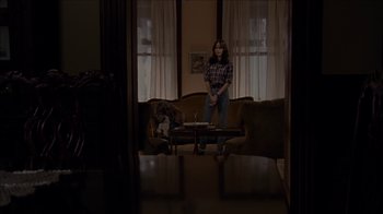 Movie still from “The House of the Devil” (2009), directed by Ti West – A woman standing in front of a coffee table in a living room; Wide shot, Over the shoulder angle