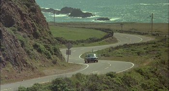 Movie still from “The Howling” (1981), directed by Joe Dante – A car driving down a curvy road near the ocean; Extreme Wide shot, High angle