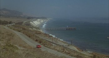 Movie still from “The Howling” (1981), directed by Joe Dante – A car driving down a road next to the ocean; Extreme Wide shot, High angle
