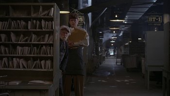 Movie still from “The Hudsucker Proxy” (1994), directed by Ethan Coen – A man standing next to a woman in a library; Medium shot, Low angle