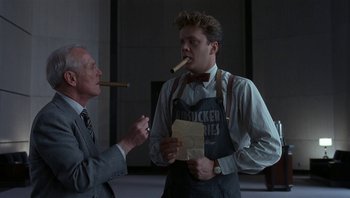 Movie still from “The Hudsucker Proxy” (1994), directed by Ethan Coen – A man smoking a cigar while another man holds a piece of paper in front of him; Medium shot, Low angle