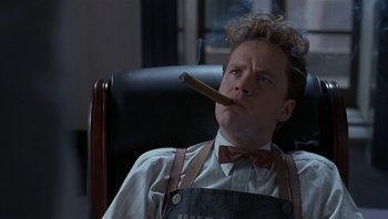 Movie still from “The Hudsucker Proxy” (1994), directed by Ethan Coen – A man sitting in a chair with a cigar in his mouth; Medium shot, Low angle