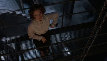 Movie still from “The Hudsucker Proxy” (1994), directed by Ethan Coen – A woman standing on a metal railing in the dark; Medium shot, Low angle