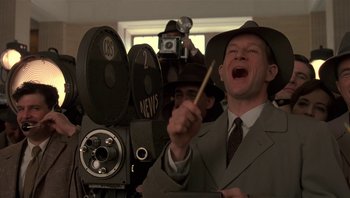 Movie still from “The Hudsucker Proxy” (1994), directed by Ethan Coen – A man in a suit and hat holding a wooden stick in front of a movie camera; Close Up shot, Low angle