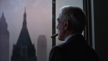 Movie still from “The Hudsucker Proxy” (1994), directed by Ethan Coen – An older man smoking a cigar in front of a window; Medium shot, Over the shoulder angle