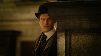Movie still from “The Illusionist” (2006), directed by Neil Burger – A man wearing a suit and a black hat standing next to a pillar; Medium shot, Low angle