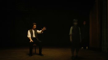 Movie still from “The Illusionist” (2006), directed by Neil Burger – A man sitting on top of a chair in front of a group of people; Wide shot, High angle