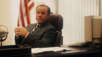 Movie still from “The Informant!” (2009), directed by Steven Soderbergh – A man sitting at a desk in front of an american flag; Medium shot, Low angle