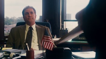 Movie still from “The Informant!” (2009), directed by Steven Soderbergh – A man sitting at a desk in front of an american flag; Medium shot, Over the shoulder angle