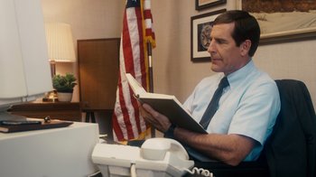 Movie still from “The Informant!” (2009), directed by Steven Soderbergh – A man sitting in front of an american flag holding an open book; Medium shot, Low angle