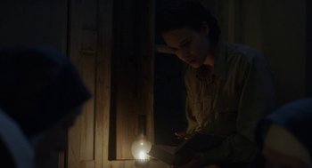 Movie still from “The Innocents” (2016), directed by Anne Fontaine – A woman reading a book in a dimly - lit room; Medium shot, Over the shoulder angle