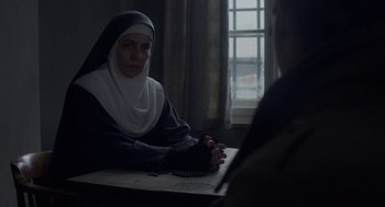 Movie still from “The Innocents” (2016), directed by Anne Fontaine – A nun sitting at a table with a man in front of a window; Medium shot, Over the shoulder angle