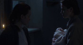 Movie still from “The Innocents” (2016), directed by Anne Fontaine – A woman holding a baby next to another woman in a dark room; Medium shot, Over the shoulder angle