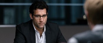 Movie still from “The International” (2009), directed by Tom Tykwer – A person wearing a suit and glasses; Close Up shot, Over the shoulder angle