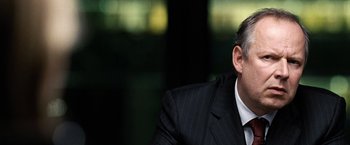 Movie still from “The International” (2009), directed by Tom Tykwer – A man wearing a suit and tie sitting in a room; Close Up shot, Over the shoulder angle