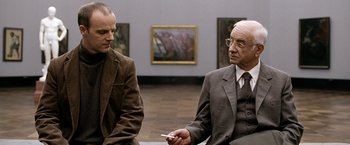Movie still from “The International” (2009), directed by Tom Tykwer – Two men are sitting in front of a painting; Medium shot, Over the shoulder angle
