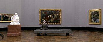 Movie still from “The International” (2009), directed by Tom Tykwer – A man sitting on a bench in front of a painting; Wide shot, Over the shoulder angle