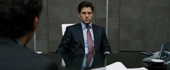 Movie still from “The International” (2009), directed by Tom Tykwer – A man sitting at a table wearing a suit and tie; Medium shot, Over the shoulder angle