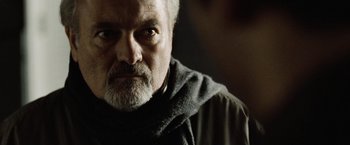 Movie still from “The International” (2009), directed by Tom Tykwer – An older man with a beard and a scarf around his neck; Close Up shot, Over the shoulder angle
