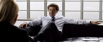 Movie still from “The International” (2009), directed by Tom Tykwer – A man sitting on top of a black couch wearing a tie; Medium shot, Low angle
