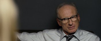 Movie still from “The International” (2009), directed by Tom Tykwer – An older man wearing glasses and a shirt and tie; Close Up shot, Over the shoulder angle