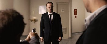 Movie still from “The International” (2009), directed by Tom Tykwer – A man in a suit and tie standing in front of a camera; Medium shot, Over the shoulder angle