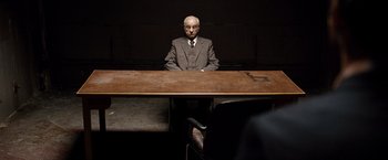 Movie still from “The International” (2009), directed by Tom Tykwer – An old man sitting at a table in a dark room; Wide shot, Over the shoulder angle