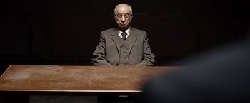 Movie still from “The International” (2009), directed by Tom Tykwer – An old man sitting at a table in a dark room; Medium shot, Over the shoulder angle