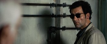 Movie still from “The International” (2009), directed by Tom Tykwer – A man wearing sunglasses standing next to pipes; Close Up shot, Over the shoulder angle