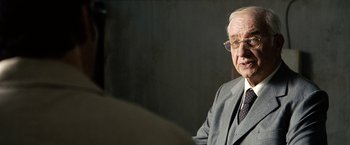 Movie still from “The International” (2009), directed by Tom Tykwer – An older man wearing a suit and tie; Close Up shot, Over the shoulder angle