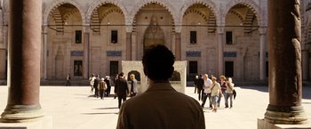 Movie still from “The International” (2009), directed by Tom Tykwer – A man standing in front of a crowd of people in front of a building; Wide shot, Over the shoulder angle