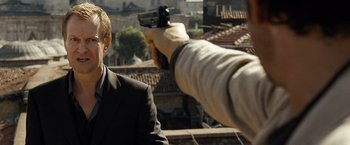 Movie still from “The International” (2009), directed by Tom Tykwer – A man in a black suit is pointing a gun at someone; Close Up shot, Over the shoulder angle