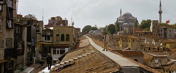 Movie still from “The International” (2009), directed by Tom Tykwer – A man standing on the side of a road in front of buildings; Extreme Wide shot, High angle