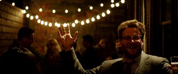 Movie still from “The Interview” (2014), directed by Evan Goldberg – A man holding up his hand in the air in front of a group of people; Medium shot, Over the shoulder angle