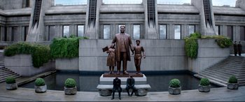 Movie still from “The Interview” (2014), directed by Evan Goldberg – A statue of a man and two children in front of a building; Wide shot, Low angle