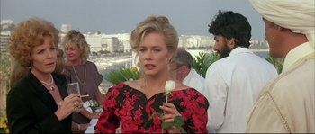 Movie still from “The Jewel of the Nile” (1985), directed by Lewis Teague – A woman holding a white rose while standing next to other people; Medium shot, Over the shoulder angle