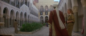 Movie still from “The Jewel of the Nile” (1985), directed by Lewis Teague – A man in a red robe and a white turban is walking down the street; Wide shot, Over the shoulder angle