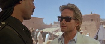 Movie still from “The Jewel of the Nile” (1985), directed by Lewis Teague – A man wearing sunglasses talking to another man in front of an outdoor scene; Close Up shot, Over the shoulder angle