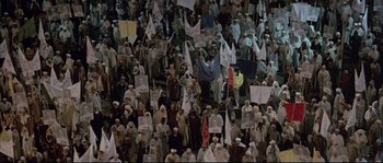 Movie still from “The Jewel of the Nile” (1985), directed by Lewis Teague – A large group of people are gathered together holding flags; Extreme Wide shot, High angle