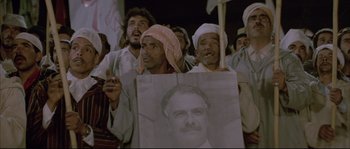Movie still from “The Jewel of the Nile” (1985), directed by Lewis Teague – A group of men holding up a sign with a picture of an old man; Medium shot, High angle